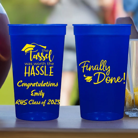 Translucent blue plastic tumbler cups customized for a graduation party. The cup on the left is personalized with "Tassel Hassle" graduation design and three lines of customer text in a semi-script font in yellow print. The cups on the right is personalized with "Finally Done!" design centered on the cup in yellow print. The cups are setting next to an ice bucket on a picnic table for a backyard graduation party