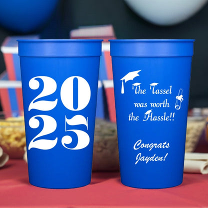 Two large blue plastic tumbler cups personalized for a 2025 graduation celebration are on a table covered with a red tablecloth with red, white and blue decorations and balloons in the background. The cup on the left displays "2025" in bold, white block style numbers. The cup in the right displays "The Tassel was worth the Hassle" in semi-script lettering with custom text "Congrats" and graduate's name in white script lettering.