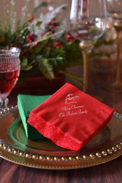 Red cocktail beverage napkins personalized with 'Reindeer' holiday design and 1 lines of custom text in gold imprint color on Christmas dinner table